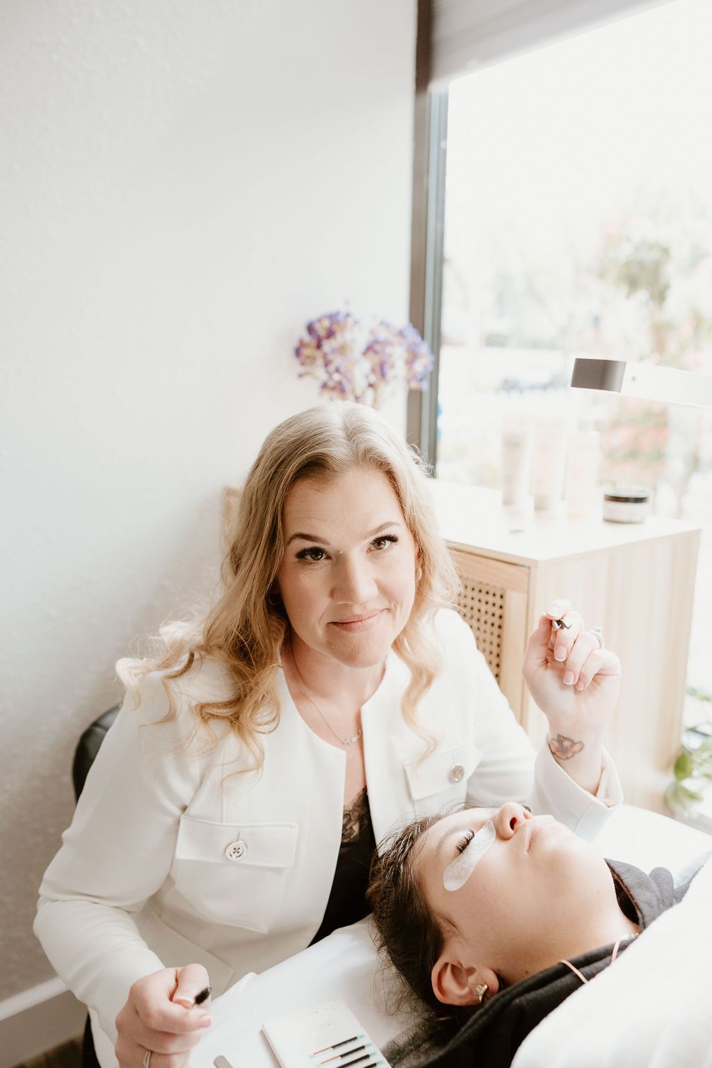 Lash artist smiling beside a client on the lash bed by a sunny window